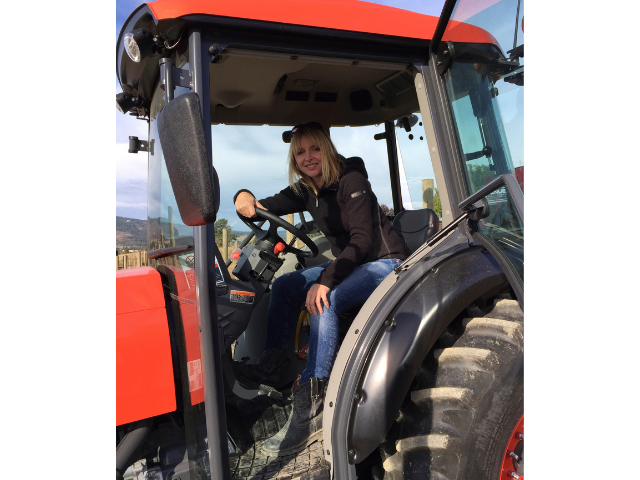 A woman with long blonde hair, wearing a black jacket and jeans is seen sitting in an orange tractor, looking at the camera smiling, with the tractor door open.
